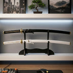Two katanas, one black and one white, displayed on a black desk stand against a modern setup with a bonsai tree and artwork from Dreams Setup.